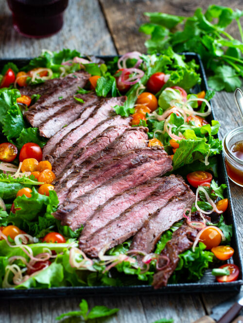 Sliced Thai beef with salad greens, tomatoes, and, fresh herbs on a metal tray with extra dressing on the side..