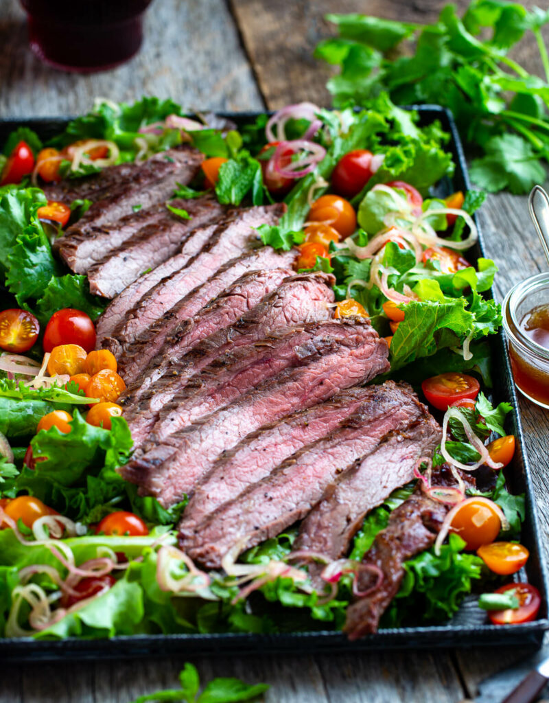 Sliced Thai beef with salad greens, tomatoes, and, fresh herbs on a metal tray with extra dressing on the side..