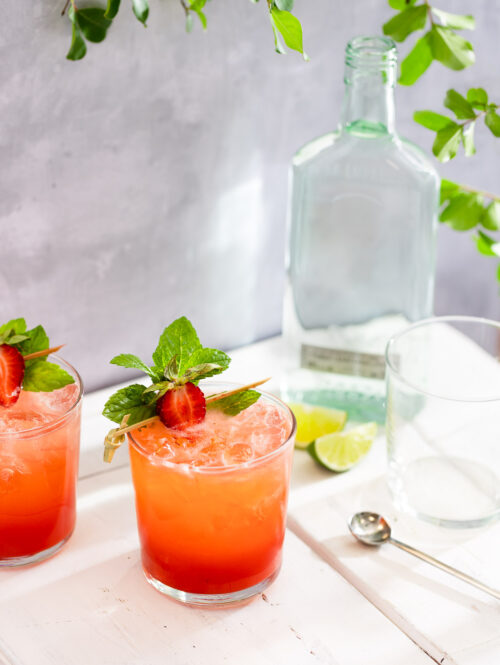 Two glasses of strawberry mojito on a white table under some leaves.