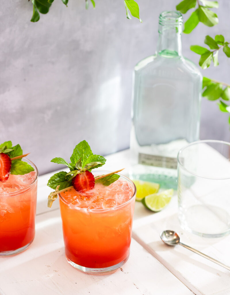 Two glasses of strawberry mojito on a white table under some leaves.