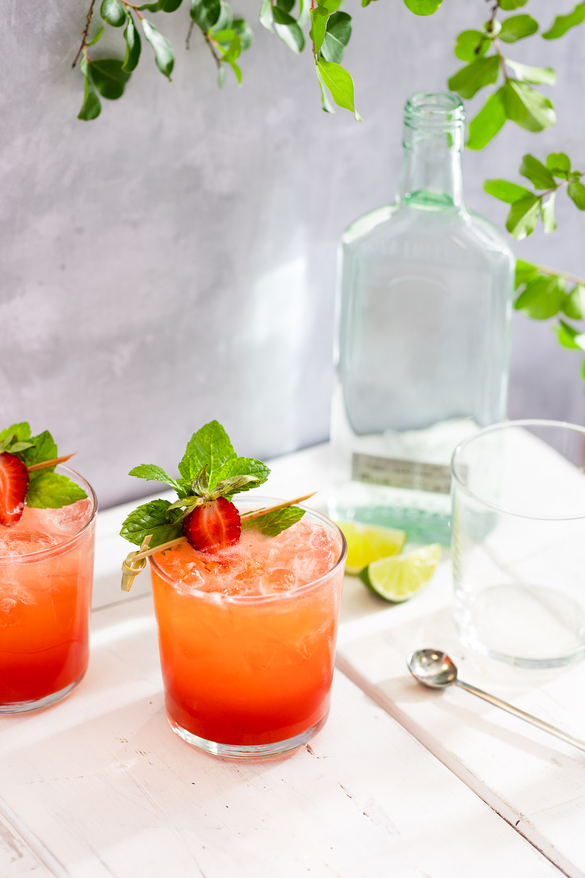 Two glasses of strawberry mojito on a white table under some leaves.