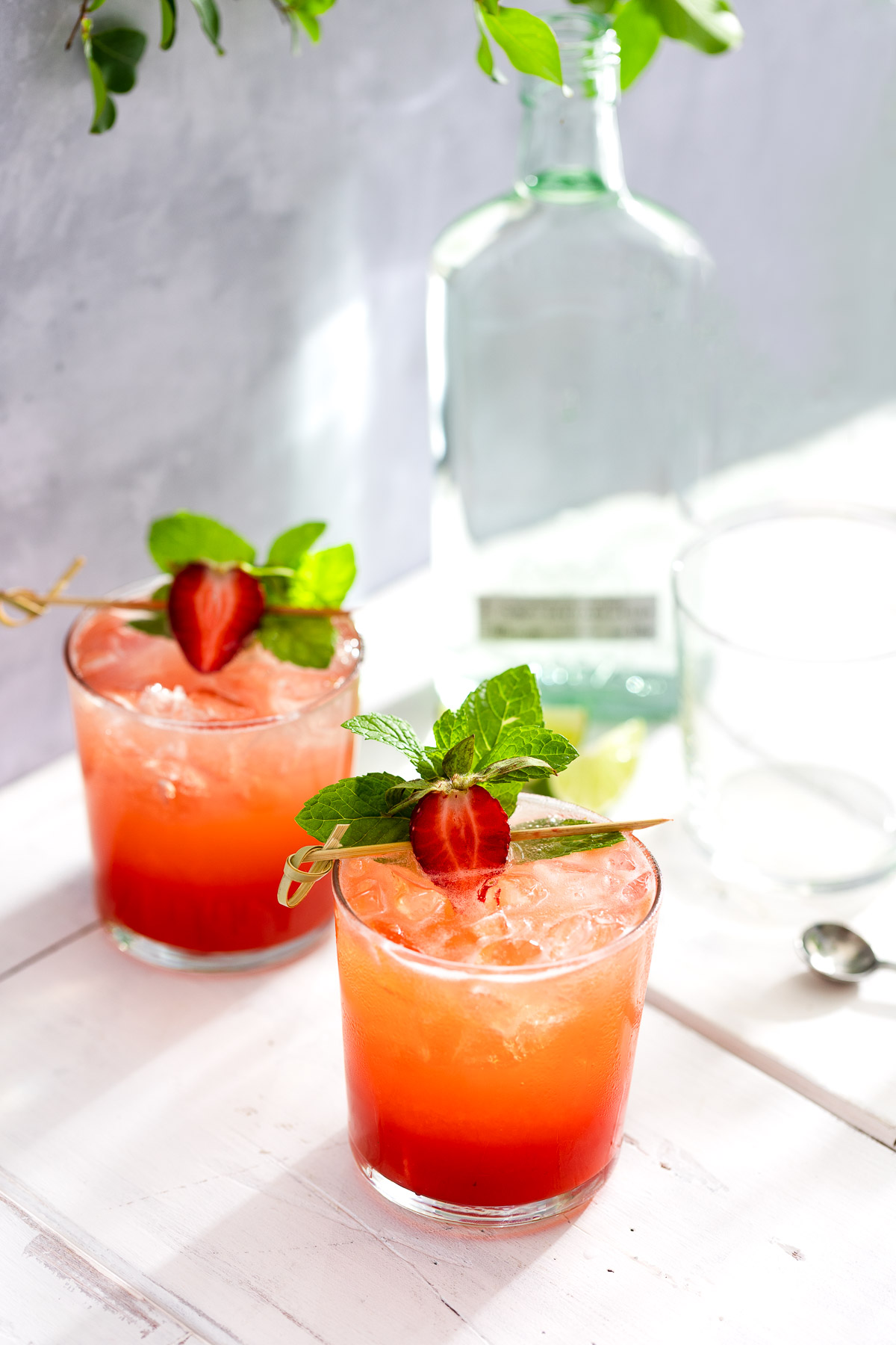Two glasses of strawberry mojito next to a bottle of rum and some lime wedges.