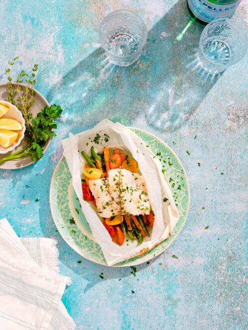 Overhead view of fish fillet and vegetables in a parchment paper packet on a green plate.