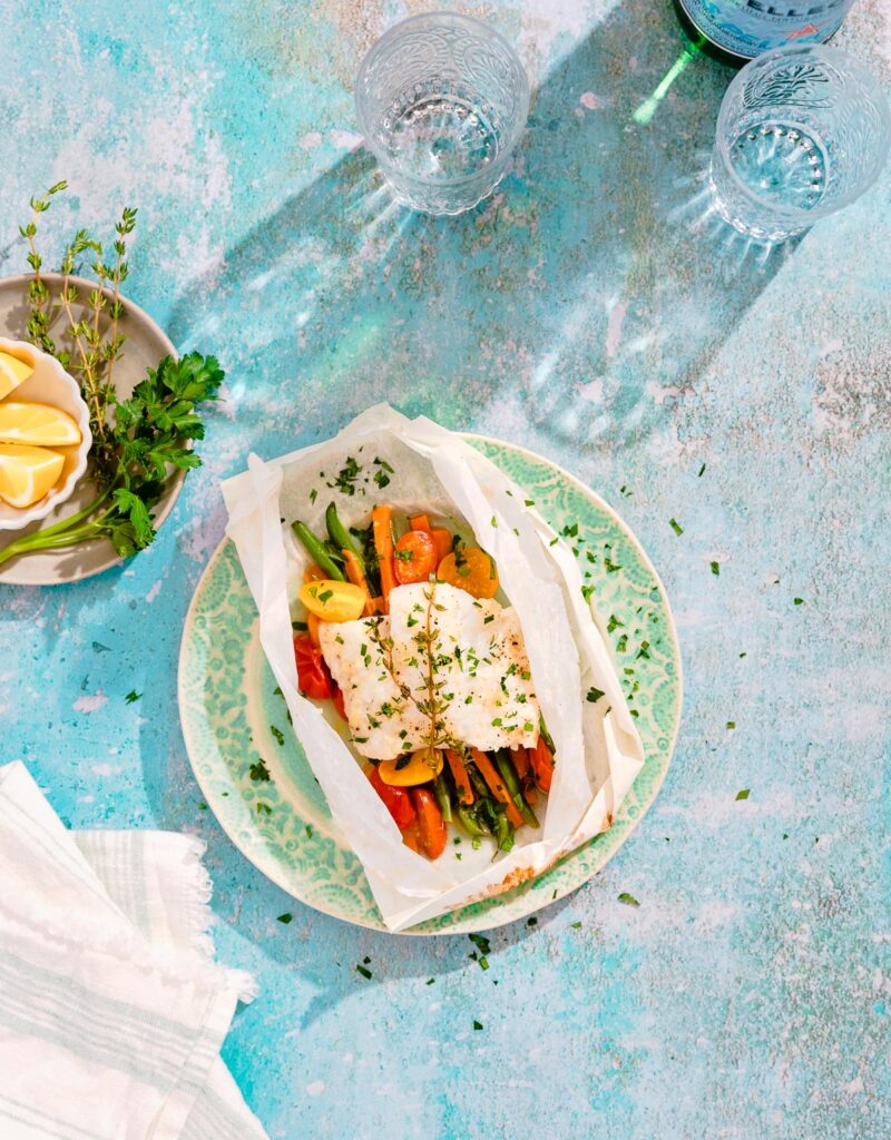 Overhead view of fish fillet and vegetables in a parchment paper packet on a green plate.