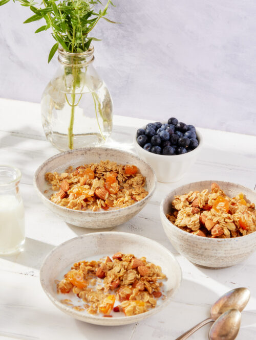 Three bowls of granola next to a bottle of milk and a bowl of blueberries.