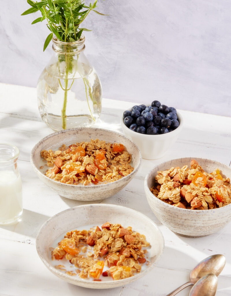 Three bowls of granola next to a bottle of milk and a bowl of blueberries.