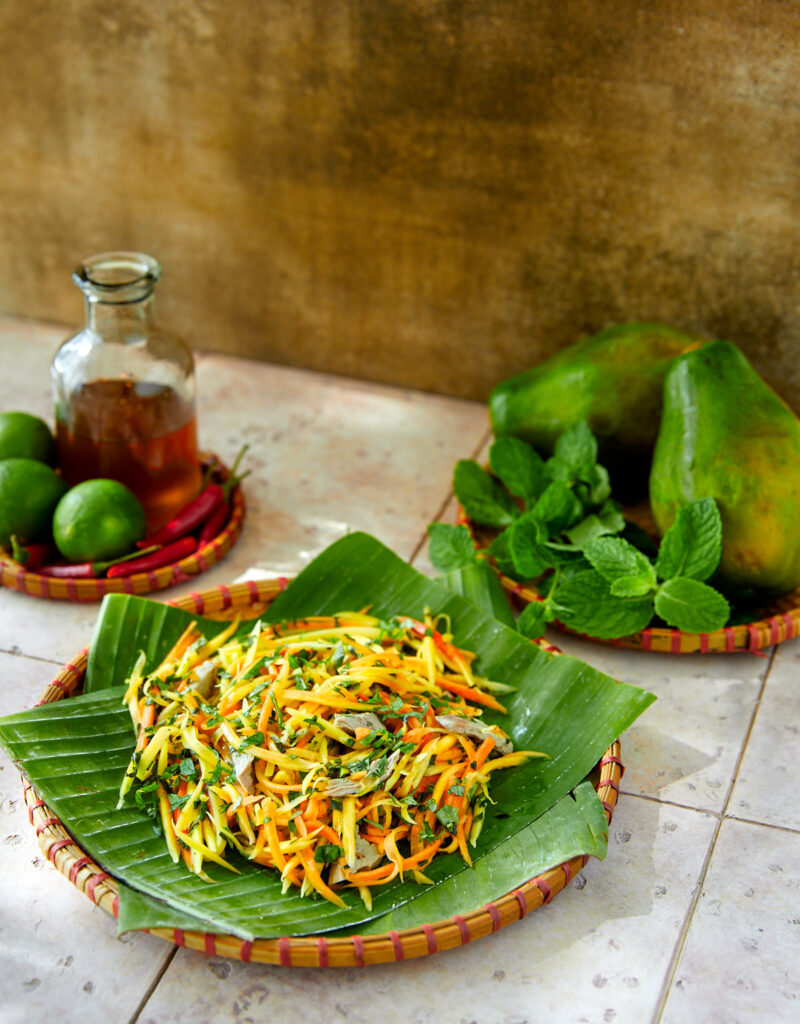 Papaya salad on some banana leaves with some mint and papaya in the background.