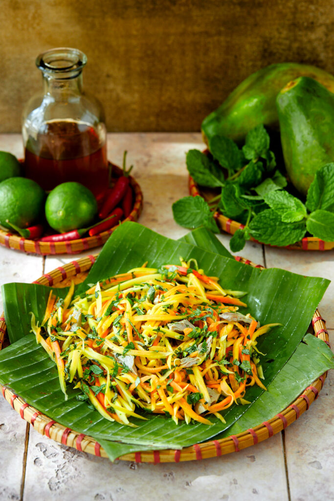 Papaya salad on some banana leaves with a jar of sauce in the background.