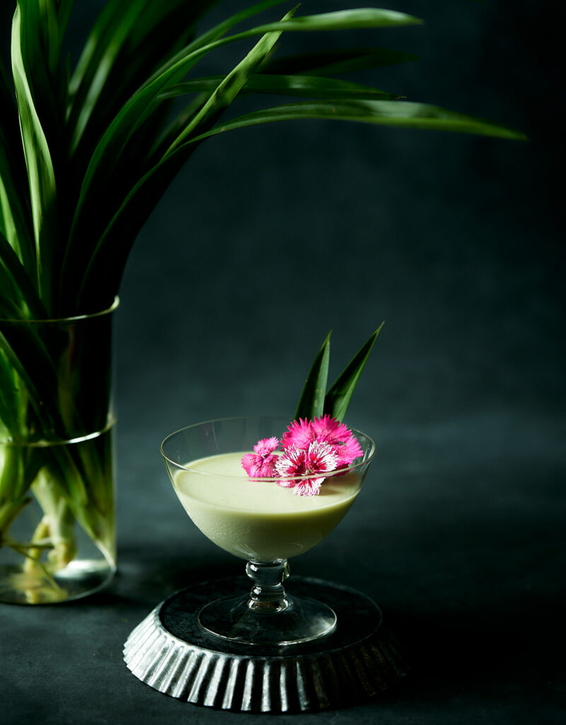 Pandan panna cotta in a short stem glass with pandan leaves in background.