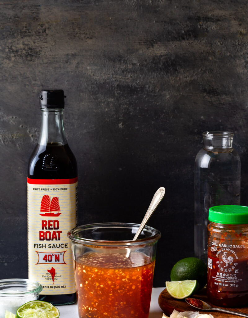 A jar of prepared Vietnamese fish sauce surrounded by a bottle of fish sauce, limes, garlic, and some chili garlic sauce.