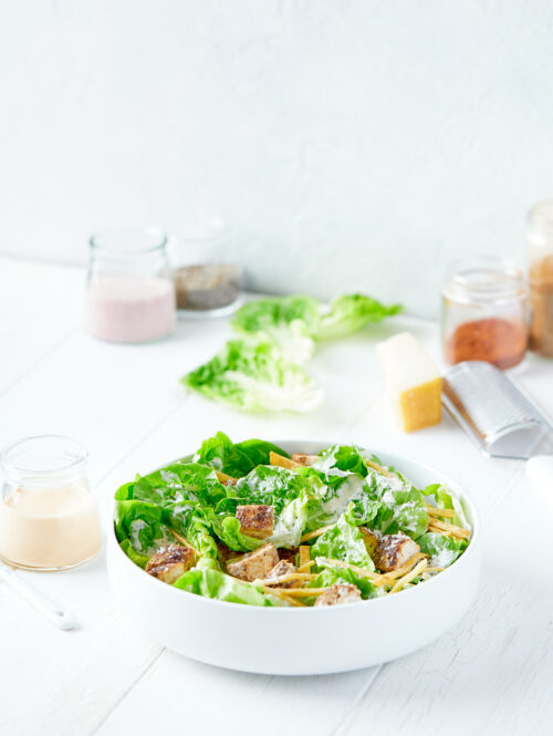 Serving bowl containing chicken caesar salad next to jar of dressing.
