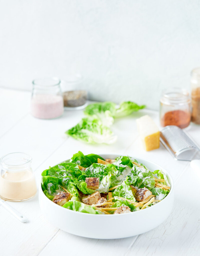 Serving bowl containing chicken caesar salad next to jar of dressing.