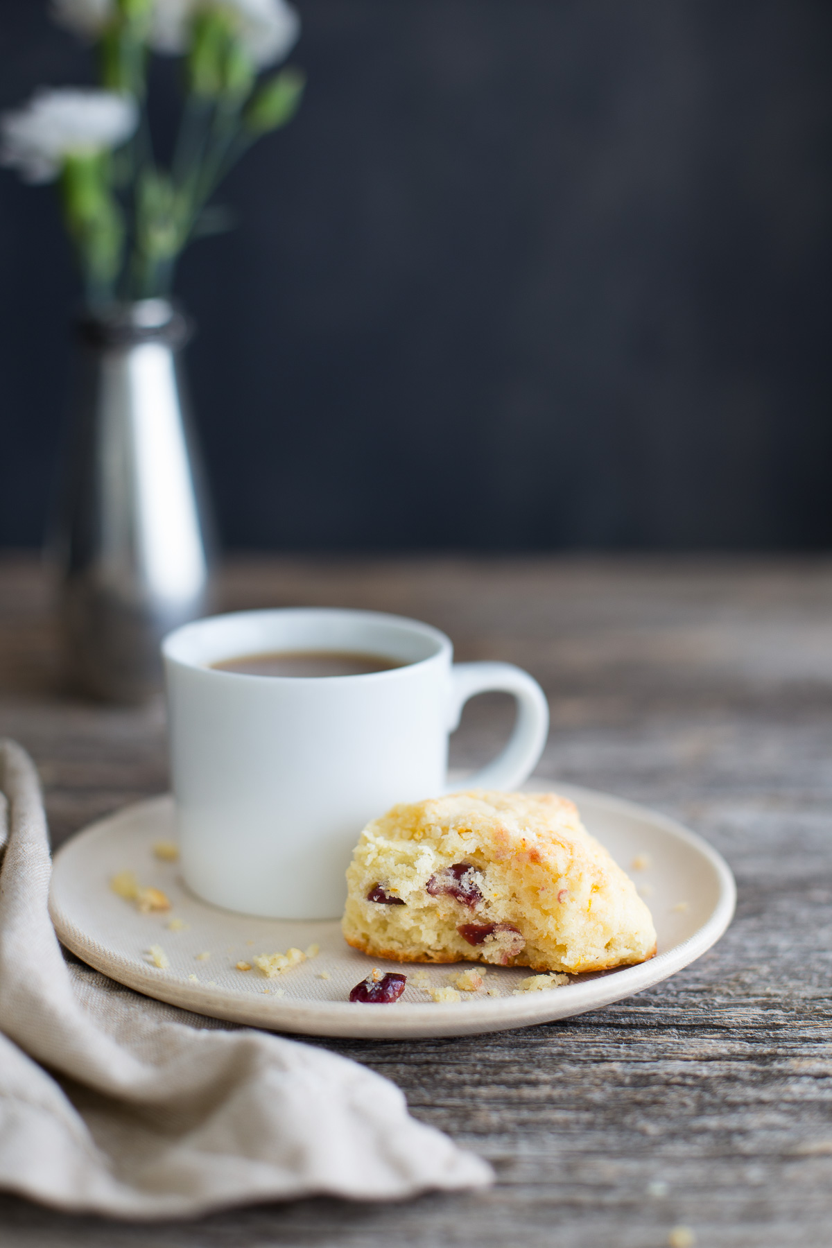 A half eaten cranberry orange scone next to a cup of tea on a white plate.