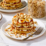A stack of pancakes layered with banana slices and macadamia nut streusel and topped with syrup.