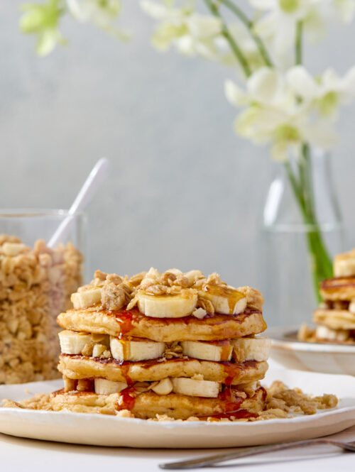 A stack of pancakes layered with banana slices and macadamia nut streusel and topped with syrup.