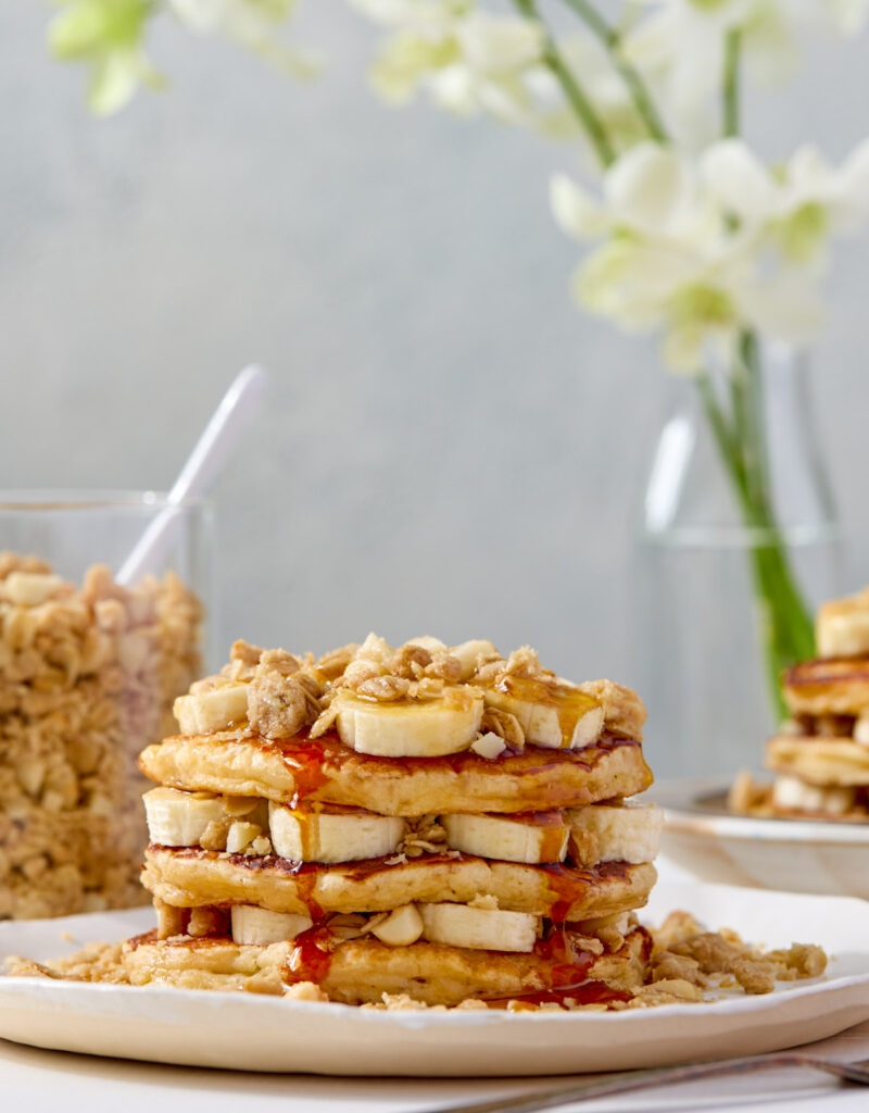 A stack of pancakes layered with banana slices and macadamia nut streusel and topped with syrup.