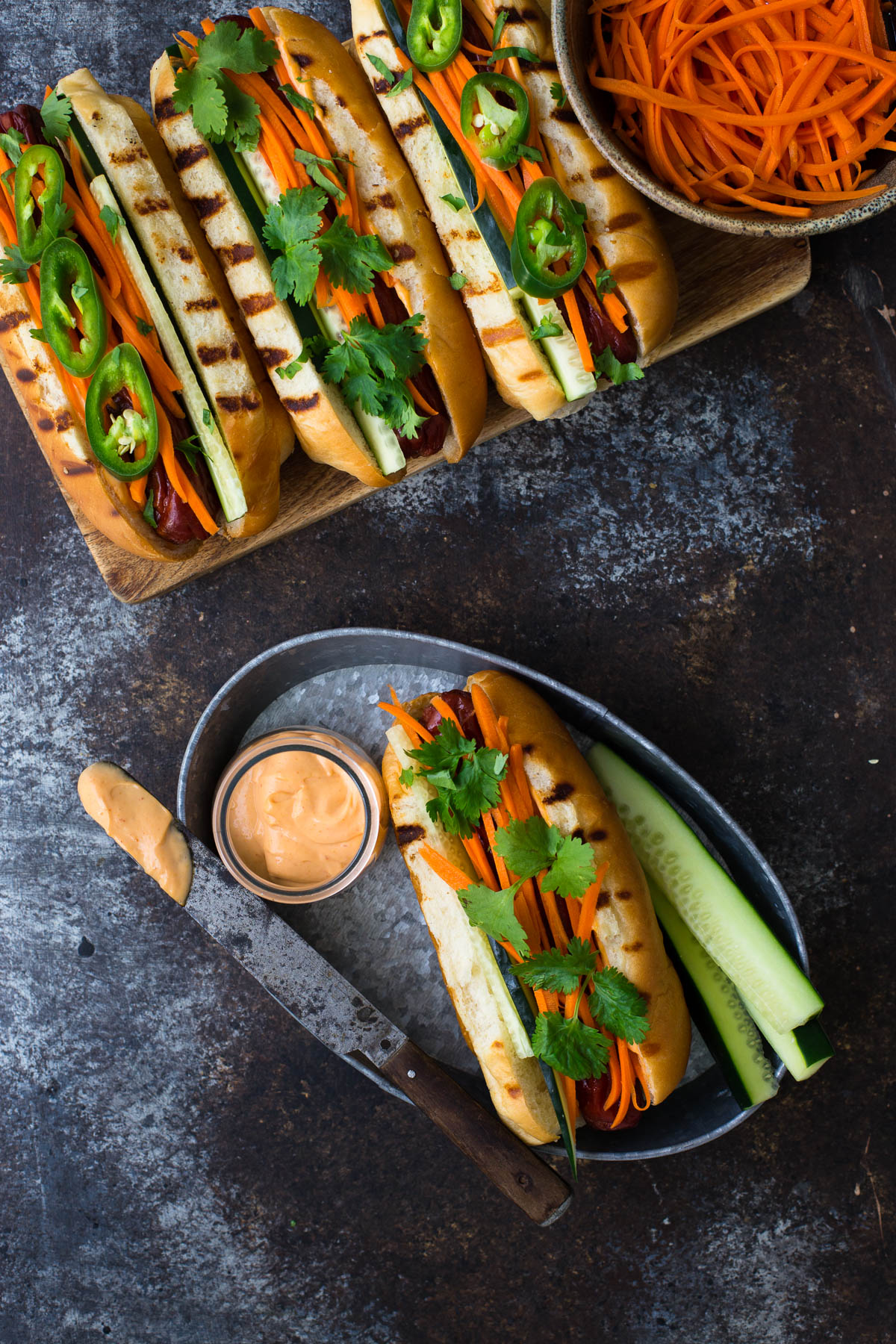 Hot dog on a metal tray topped with pickled carrots, cilantro, and sriracha mayo.