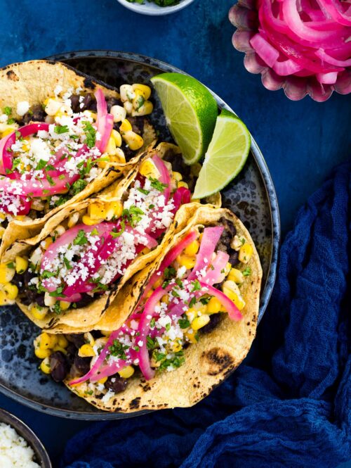 Black bean and elote tacos garnished with queso fresco, chopped cilantro, and pickled onions on a black plate.