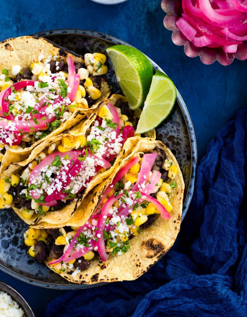 Black bean and elote tacos garnished with queso fresco, chopped cilantro, and pickled onions on a black plate.