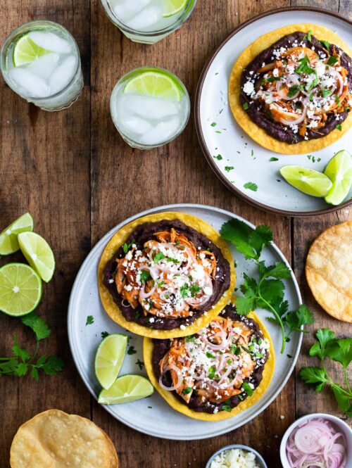 Two plates of tostadas topped with refried beans, shredded chicken, pickled shallots, and cotija cheese.