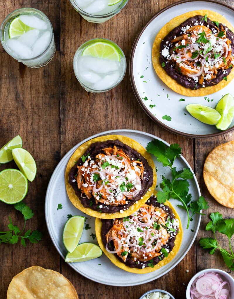 Two plates of tostadas topped with refried beans, shredded chicken, pickled shallots, and cotija cheese.