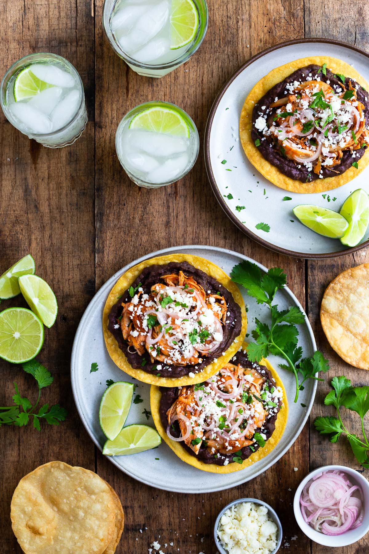 Two plates of tostadas topped with refried beans, shredded chicken, pickled shallots, and cotija cheese.