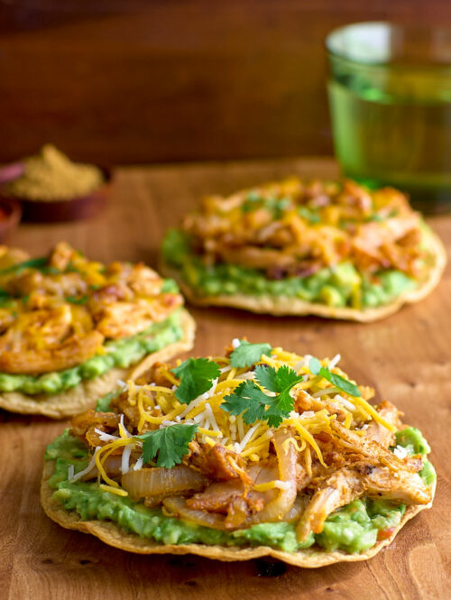A tostada topped with guacamole, sautéed onions, spiced chicken, grated cheese, and cilantro.