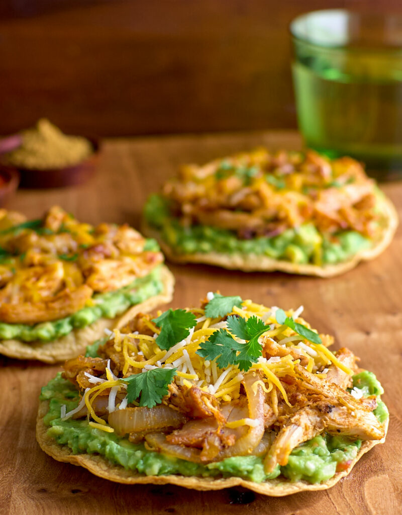 A tostada topped with guacamole, sautéed onions, spiced chicken, grated cheese, and cilantro.