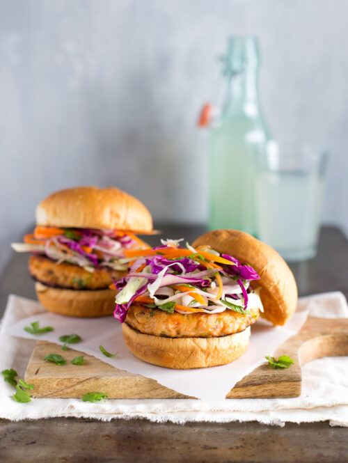 Two salmon burgers topped with cabbage slaw on a wooden board.
