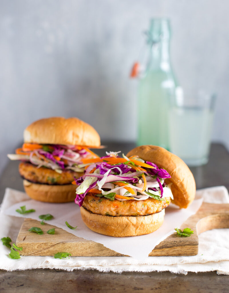 Two salmon burgers topped with cabbage slaw on a wooden board.
