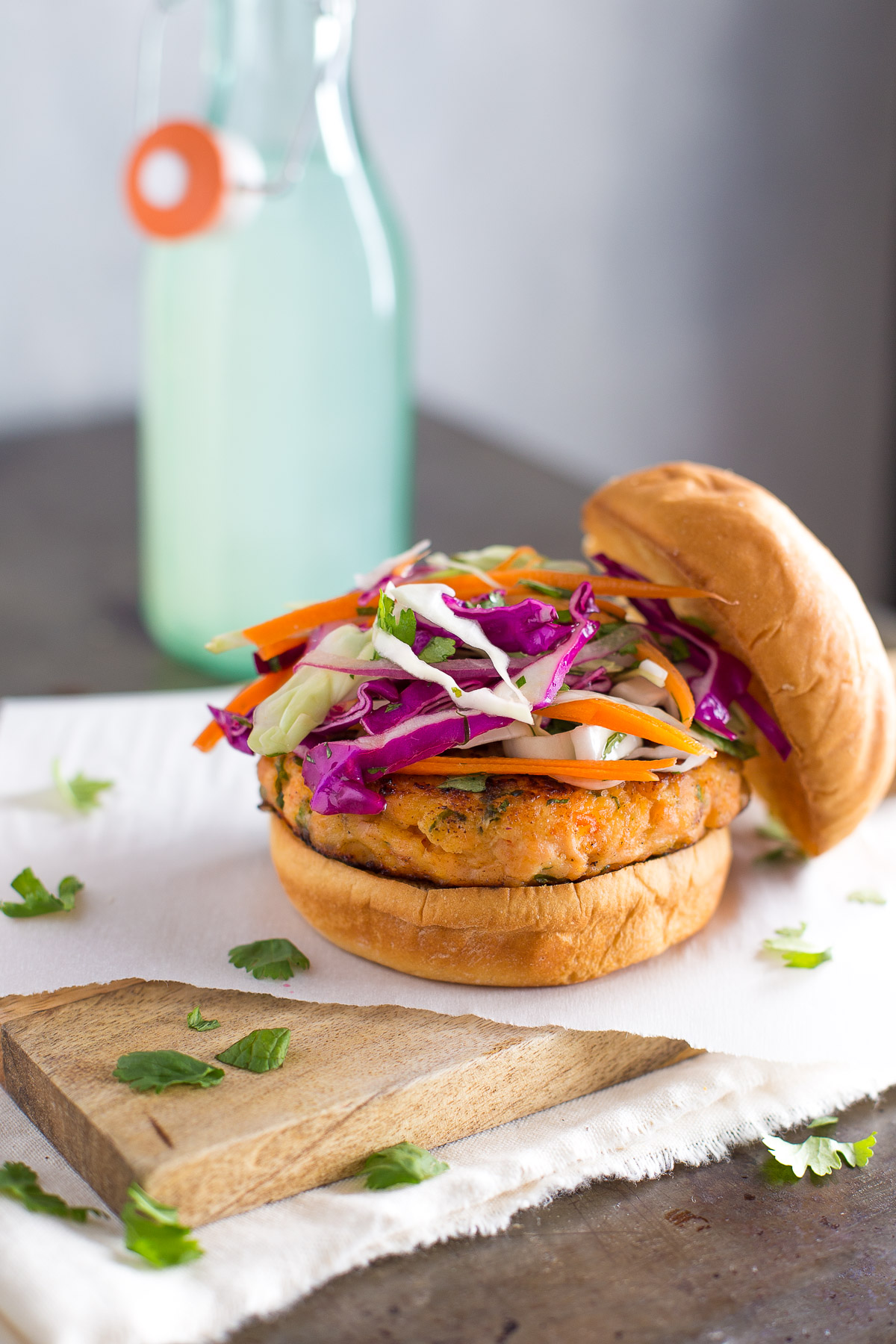A salmon burger topped with cabbage slaw on a wooden board with a bottle of lemonade in the background.