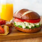 An egg salad sandwich on a pretzel roll served with chips and a glass of orange juice.