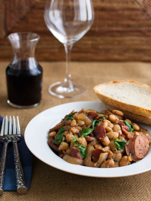 White bean stew in a white bowl next to 2 forks and a glass of red wine.
