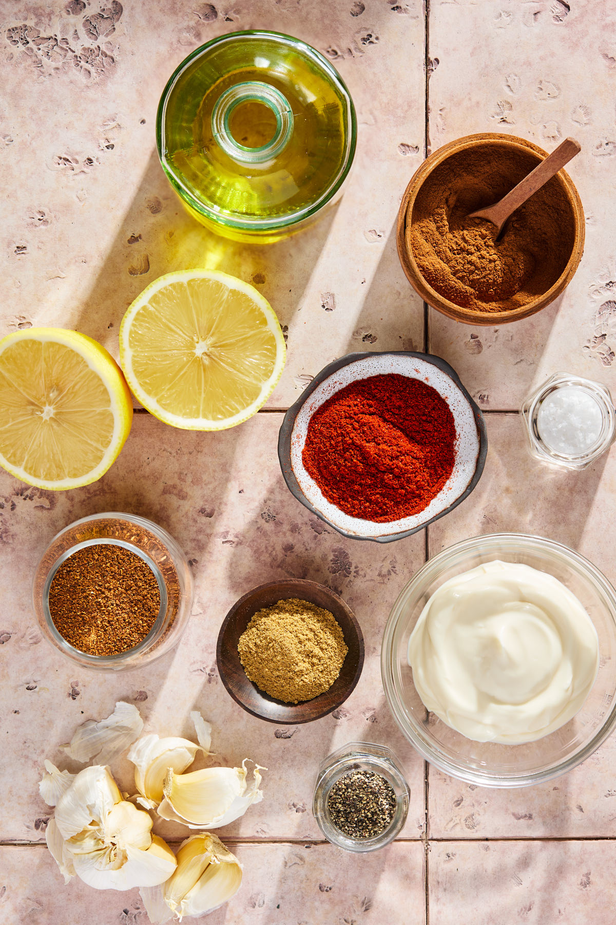 Bottle of olive oil, jars of spices, garlic cloves, and lemon on a tiled countertop.