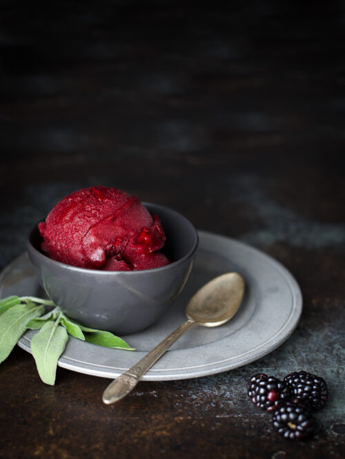 A scoop of blackberry sorbet in a small gray bowl garnished with a sprig of sage.