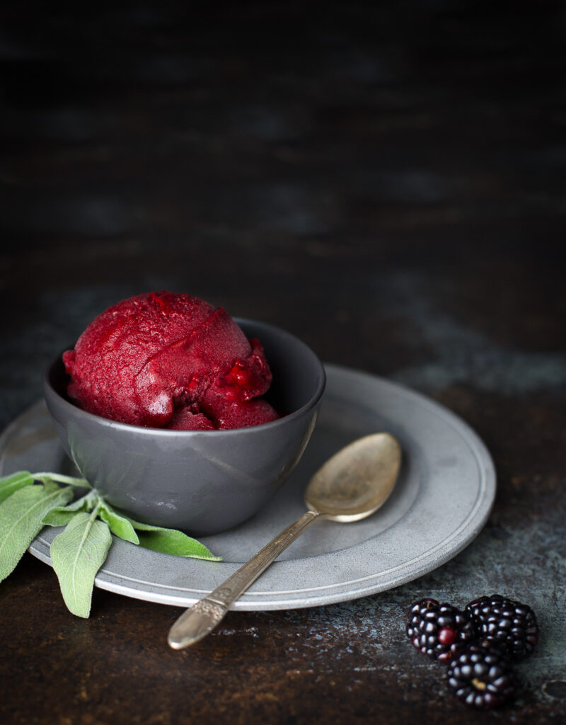 A scoop of blackberry sorbet in a small gray bowl garnished with a sprig of sage.