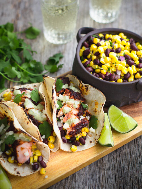 Salmon tacos next to some lime wedges and a bowl of black bean and corn relish.