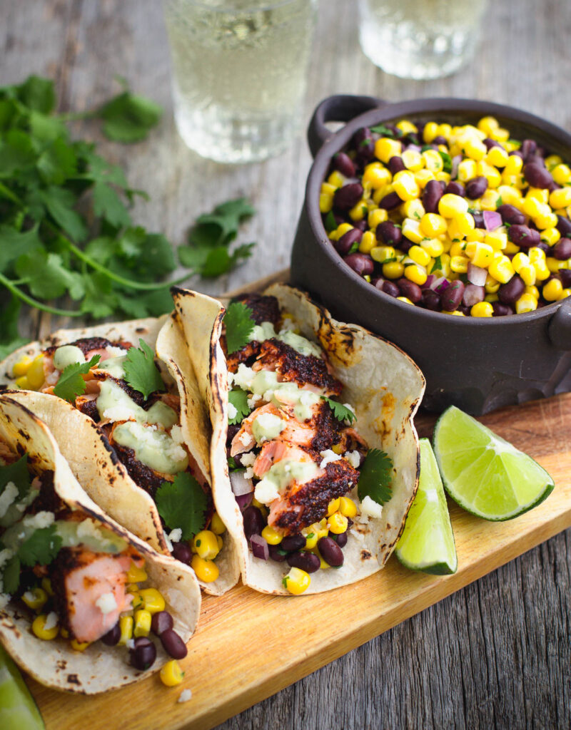 Salmon tacos next to some lime wedges and a bowl of black bean and corn relish.