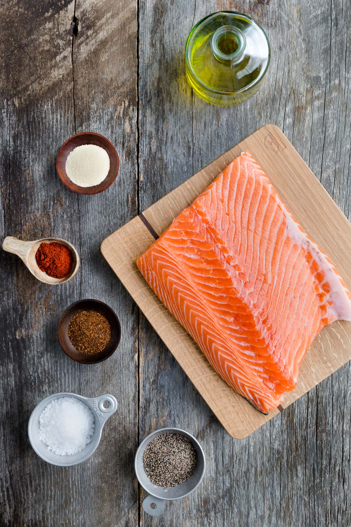 A piece of salmon on a cutting board surrounded by bowls of spices and a bottle of olive oil.