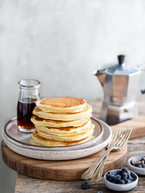 A stack of pancakes next to a bottle of syrup and small bowls of blueberries and chocolate chips.