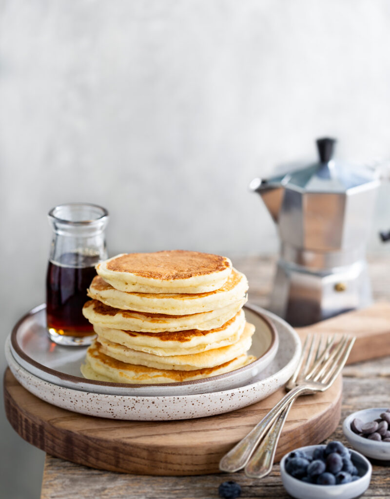 A stack of pancakes next to a bottle of syrup and small bowls of blueberries and chocolate chips.