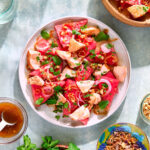 Plate of watermelon salad surrounded by plates of fresh herbs, peanuts, and fried shallots.
