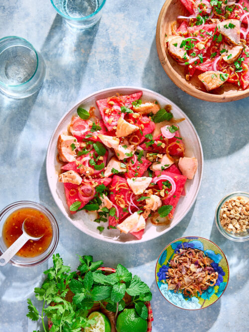 Two plates of watermelon salad surrounded by plates of fresh herbs, peanuts, and fried shallots.