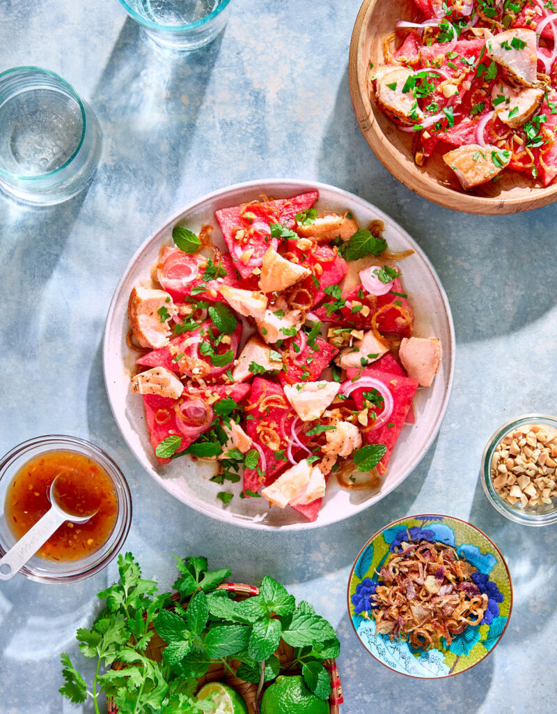 Two plates of watermelon salad surrounded by plates of fresh herbs, peanuts, and fried shallots.