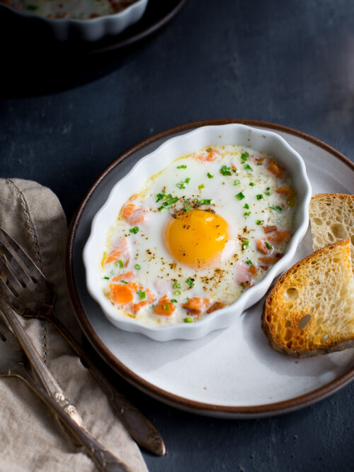 A baked egg topped with chopped chives in a white ramekin next to some toast.