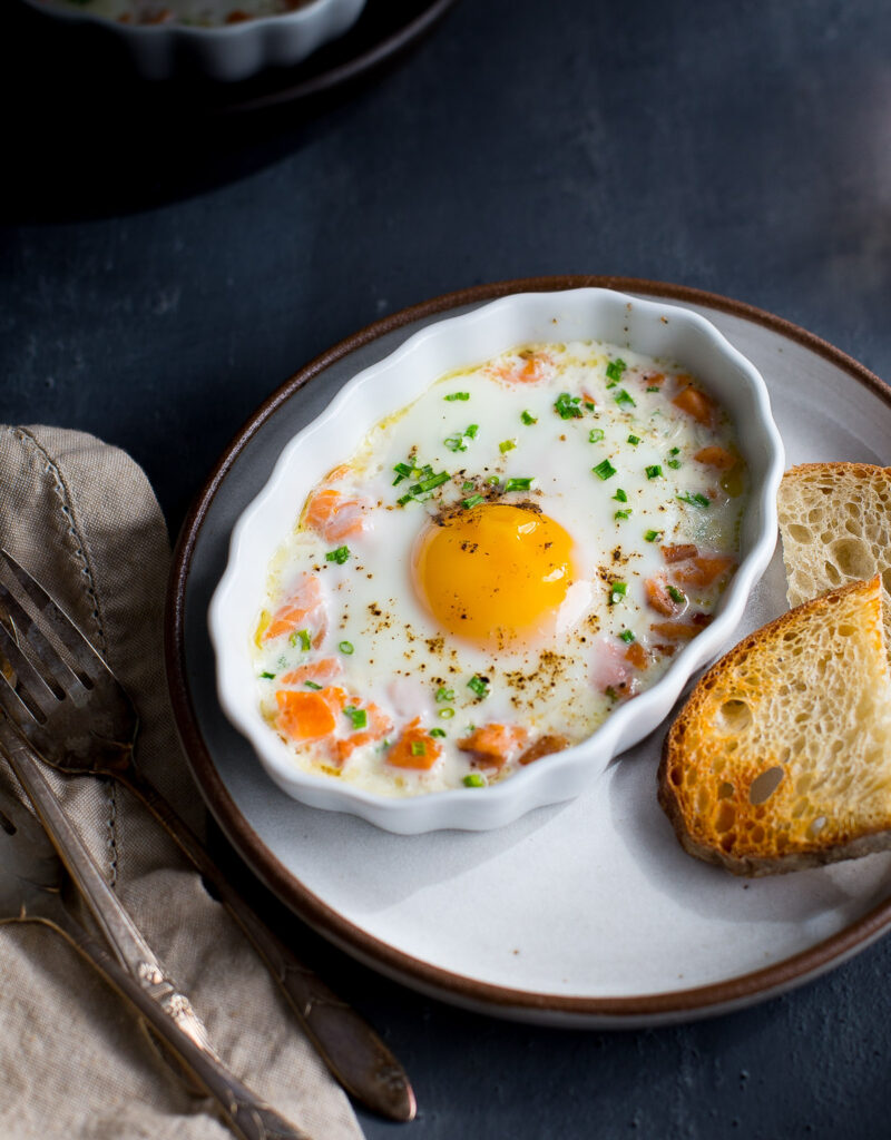 A baked egg topped with chopped chives in a white ramekin next to some toast.