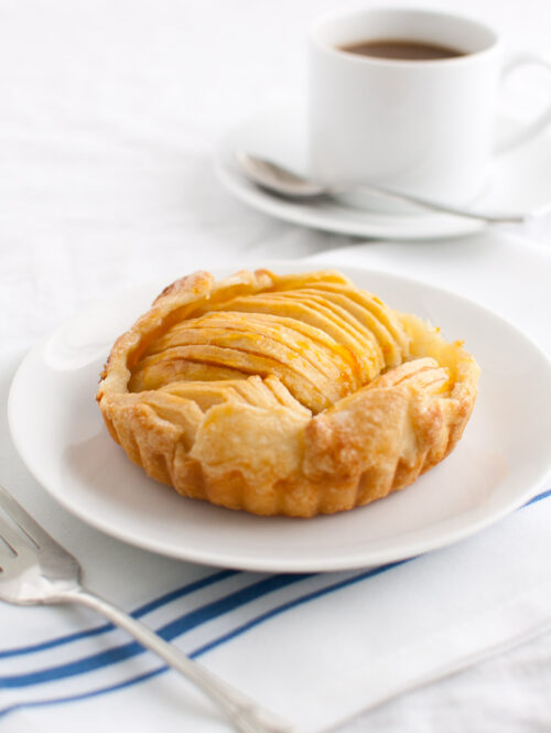 Apple tartlet on a white plate with a cup of coffee in the background.