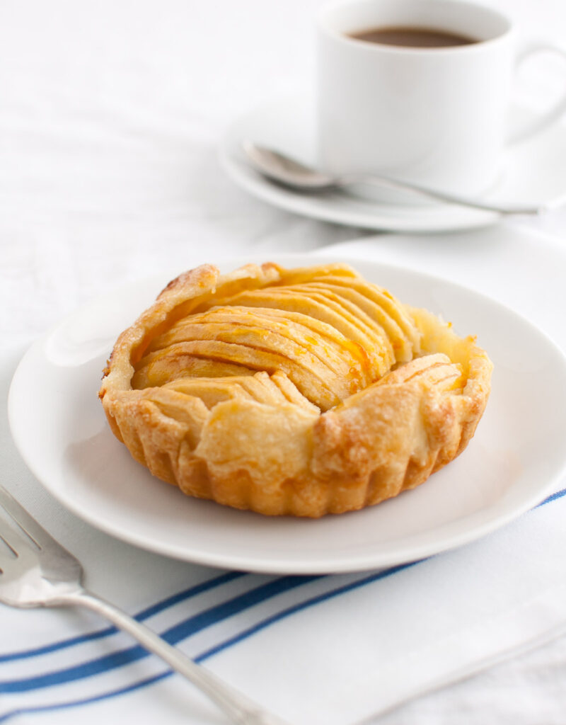 Apple tartlet on a white plate with a cup of coffee in the background.