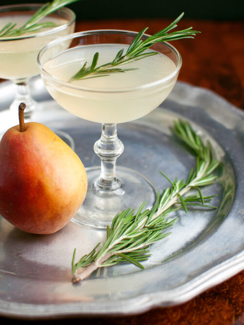 A pear-rosemary cocktail in a coupe glass garnished with a sprig of rosemary.