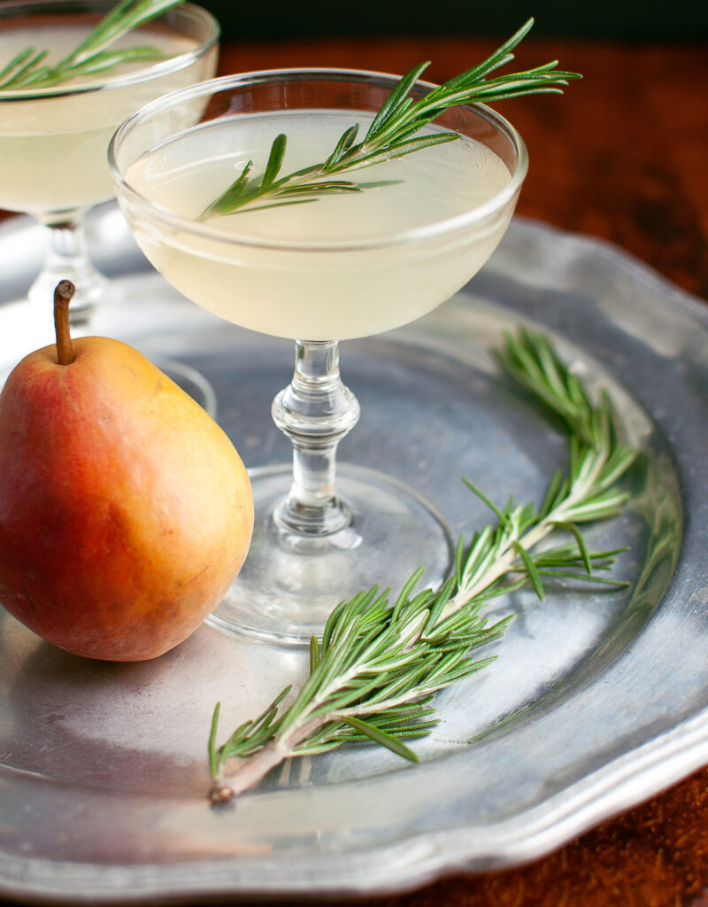 A pear-rosemary cocktail in a coupe glass garnished with a sprig of rosemary.